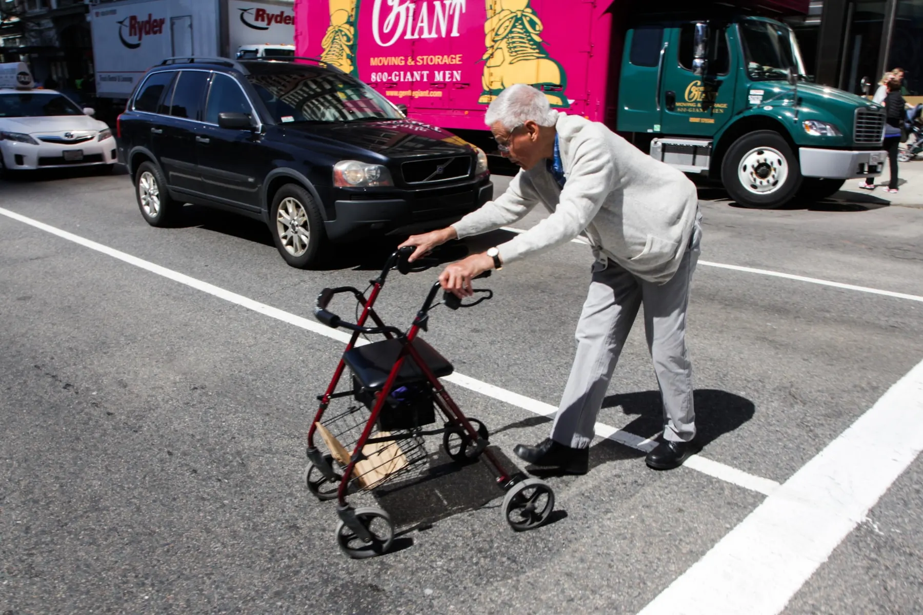 Man in pale grey reaches for a walker rolling away from him in the road