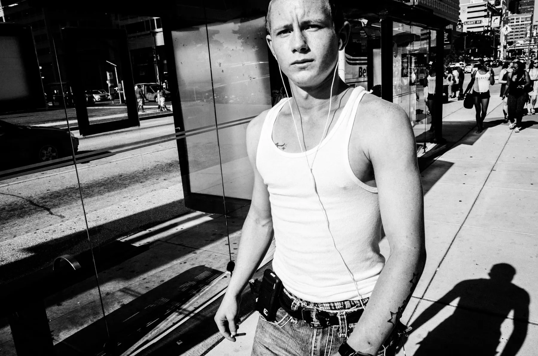 Young man in a white vest and earphones stands at a bus shelter, looking back