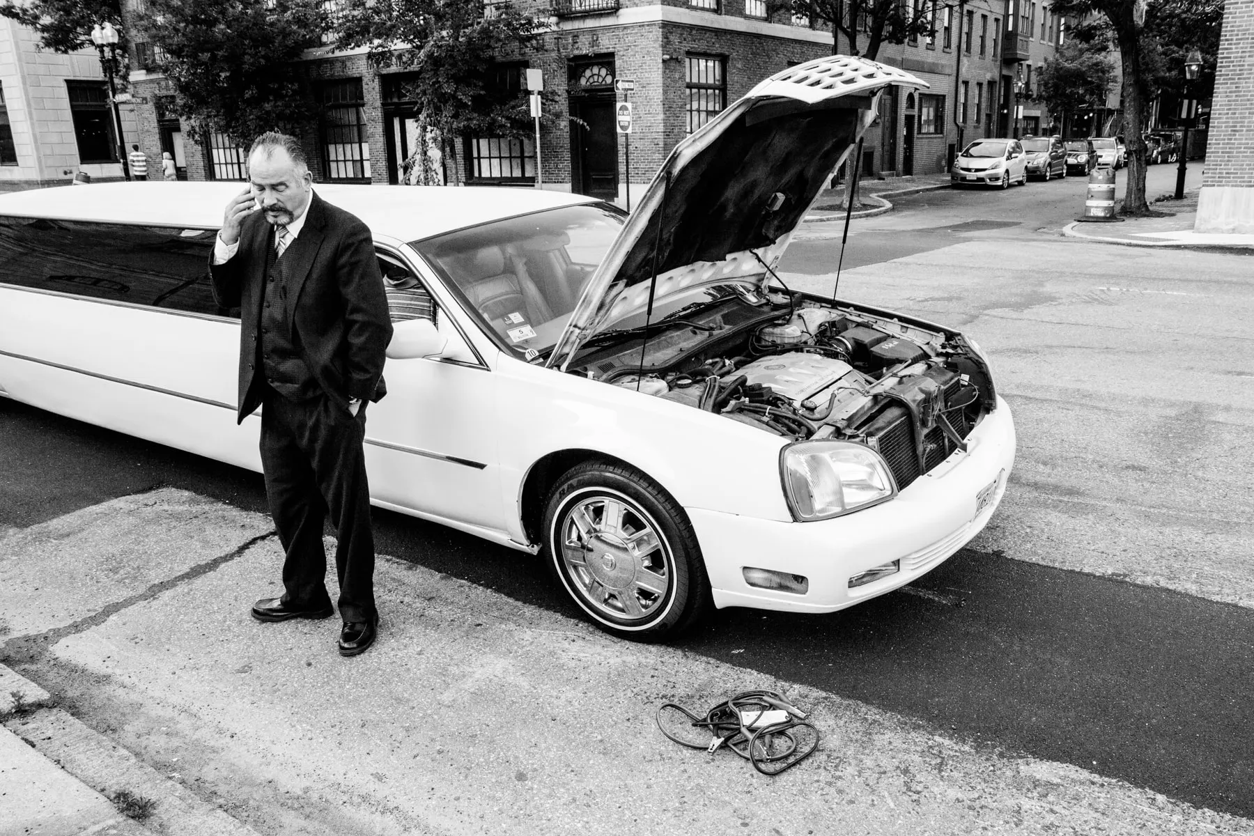 Man in a suit stands by a white sedan with its hood up, hand over his face
