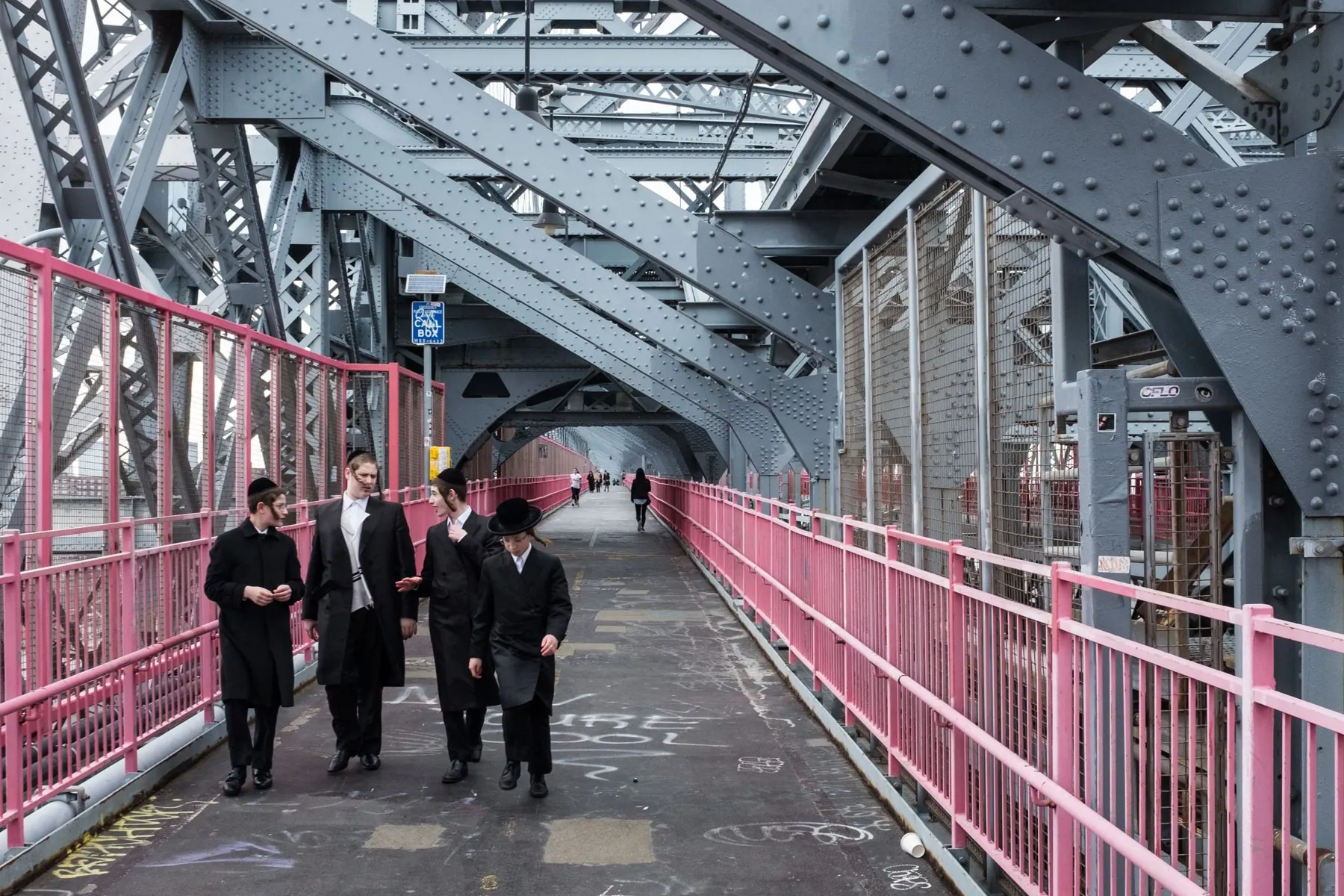 Group in black coats and hats walk a pink-railed pedestrian bridge