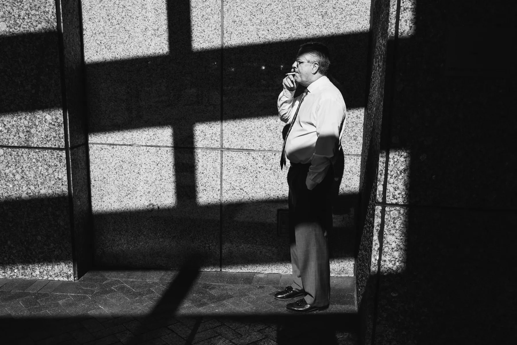 Man in shirtsleeves smokes against a wall lit by a grid of window light