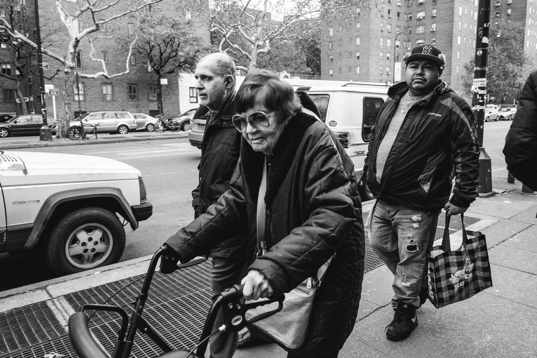 Older woman pushes a walker up a sidewalk, two men close behind her