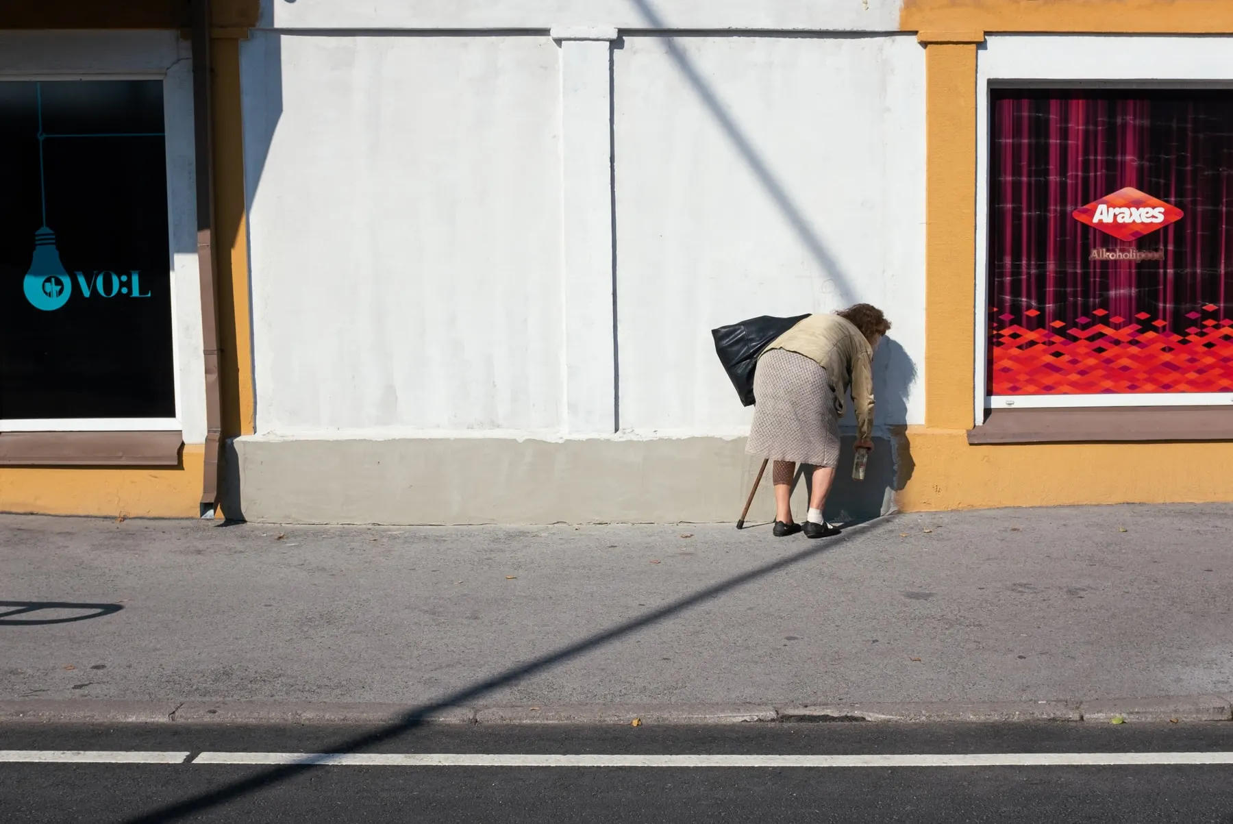 Woman with a cane bends toward a wall on a sunlit empty street