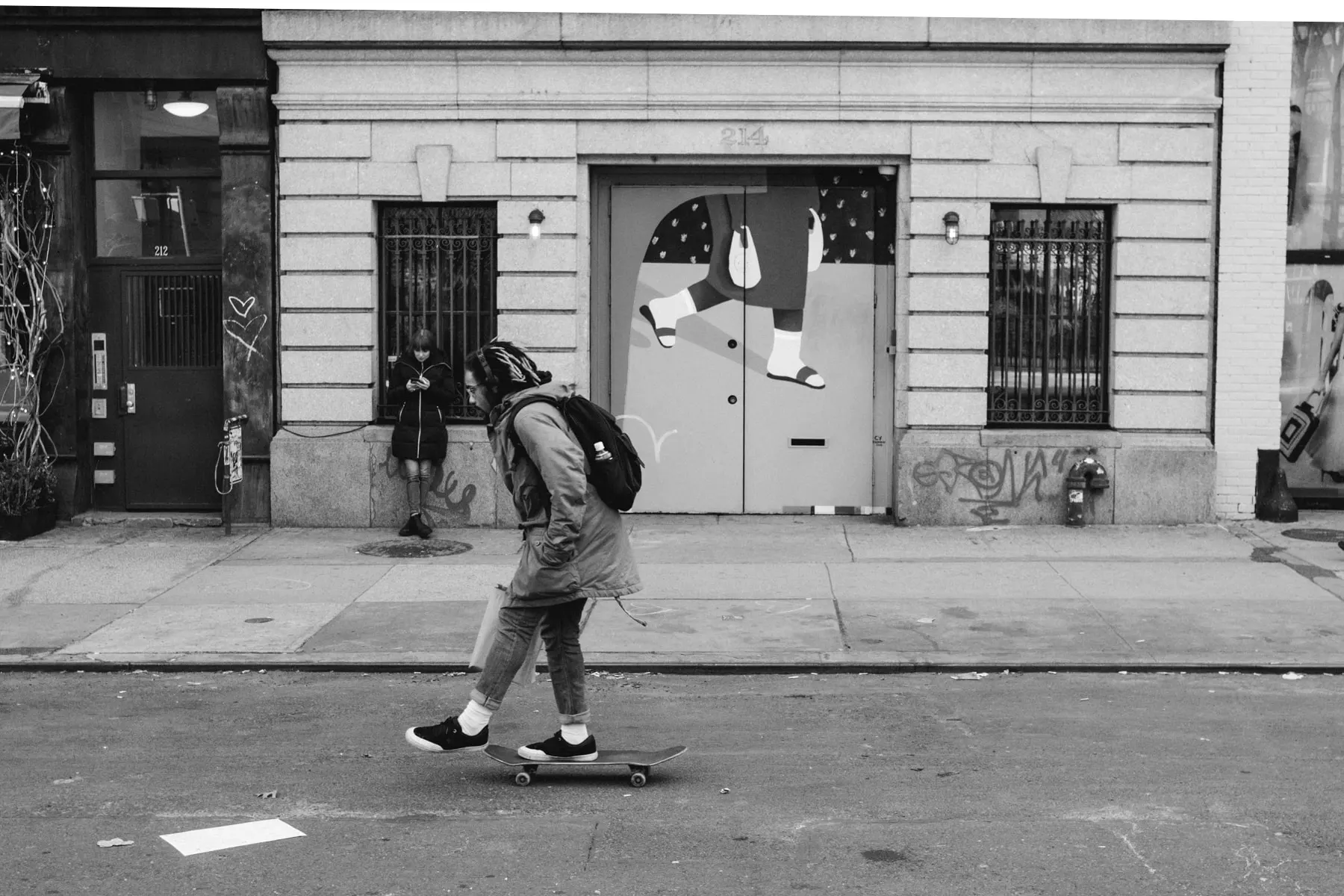 Skateboarder rolls past a shuttered storefront painted with dangling legs