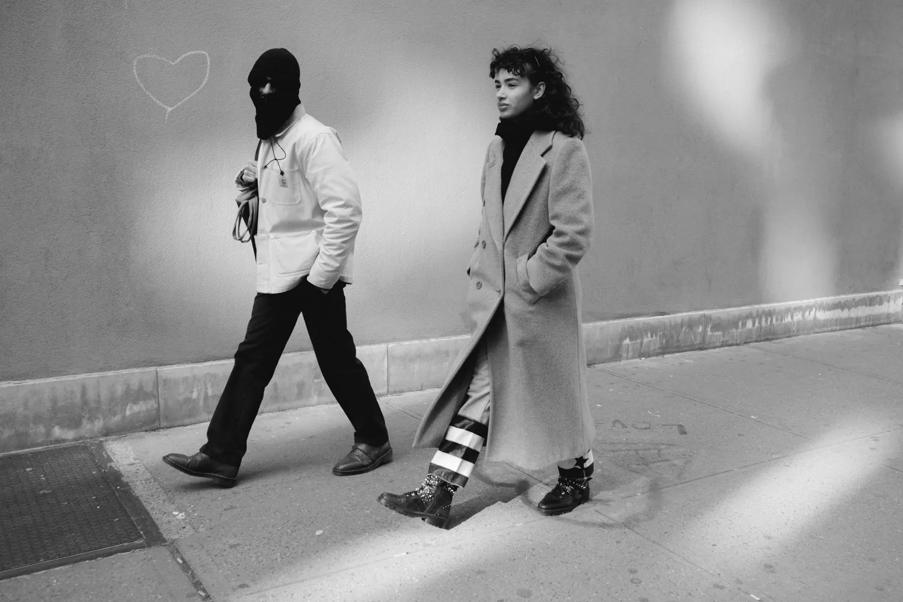 Man in a balaclava and a woman in a long coat pass each other on the pavement
