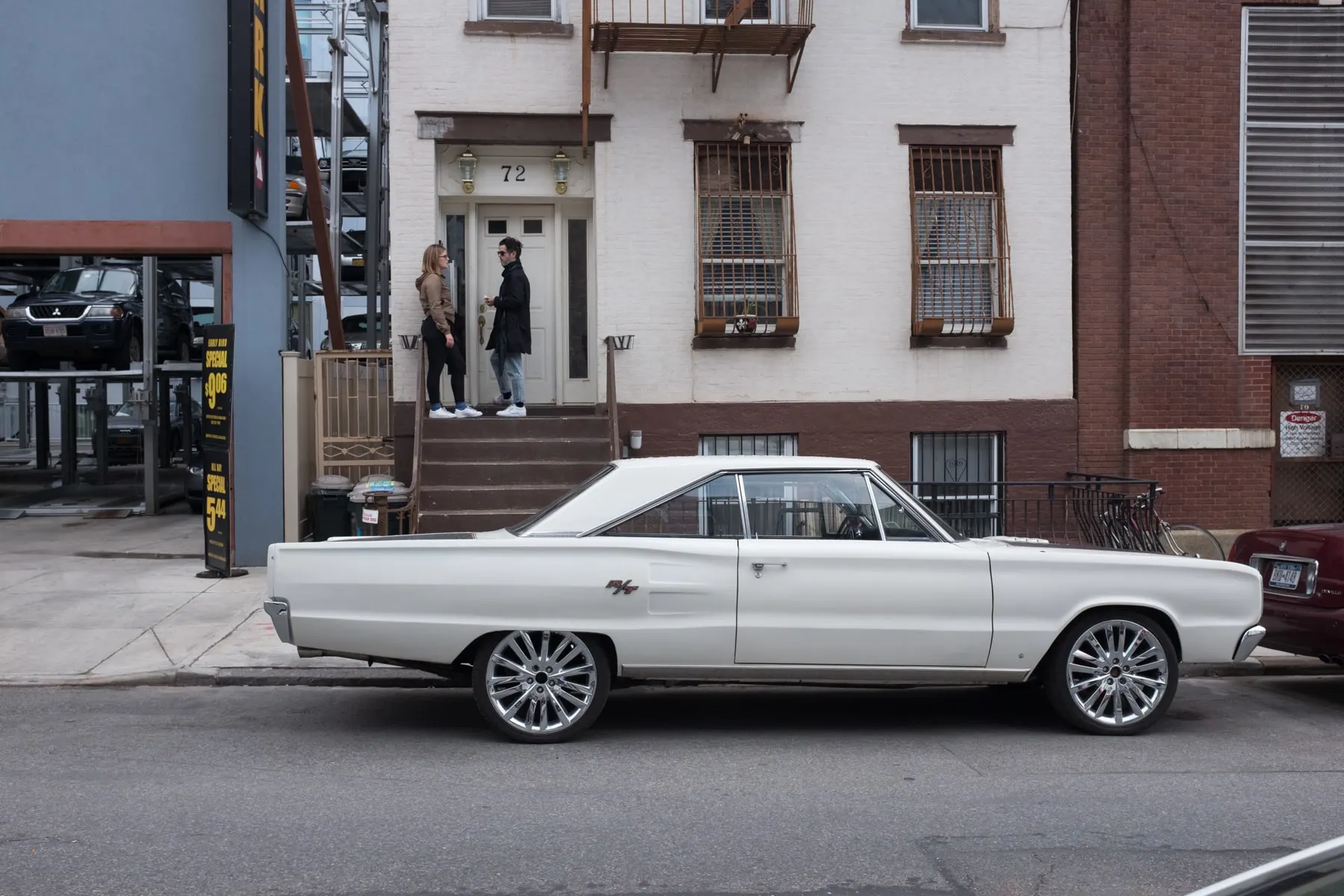 White muscle car parked below a stoop where two people stand
